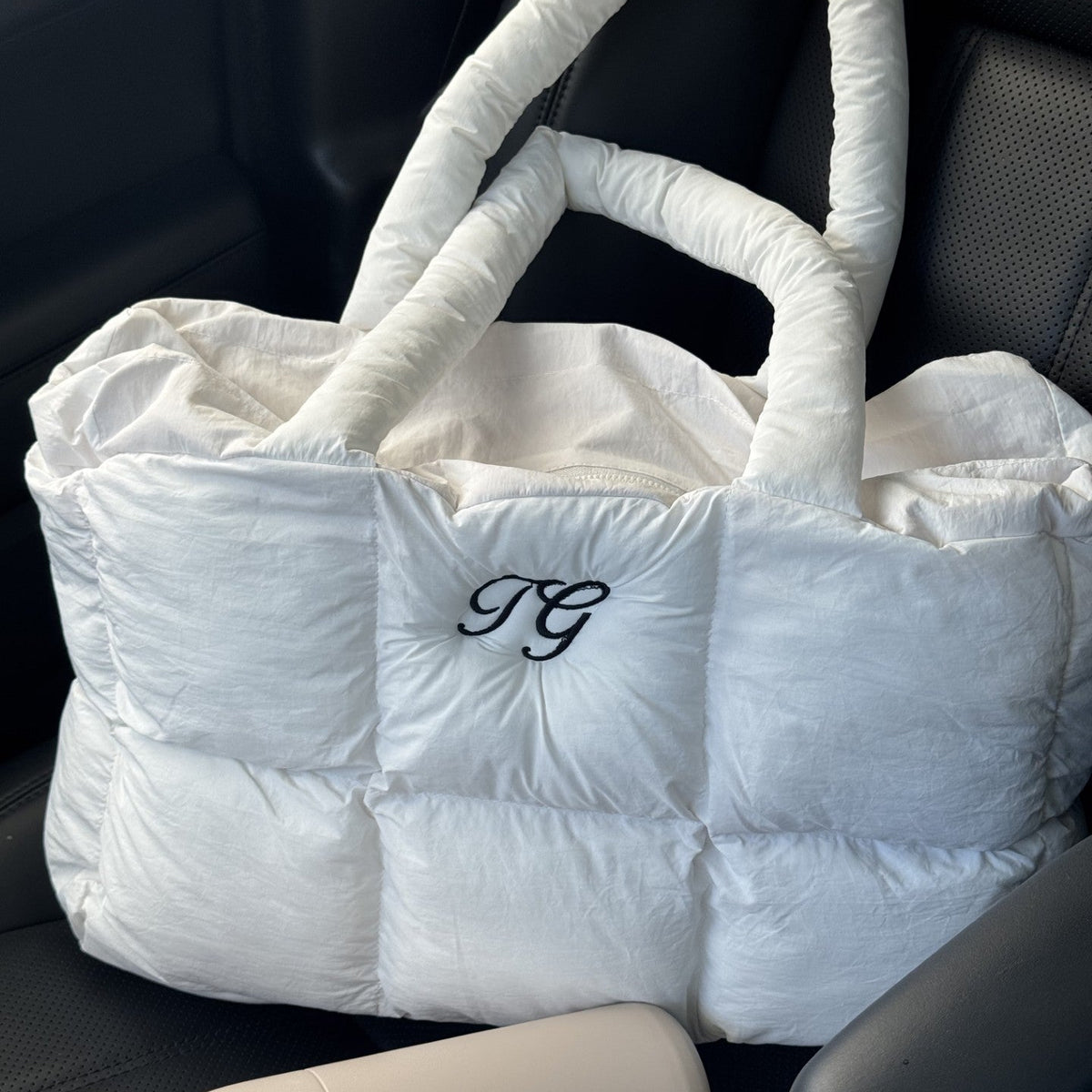 A white puffer tote bag with padded handles and a black embroidered monogram 'JG' on the front, resting on a black car seat next to a beige bottle.