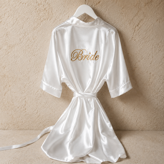 A white satin bridal robe hanging on a white hanger against a textured beige wall. The back of the robe features the word 'Bride' embroidered in gold script.