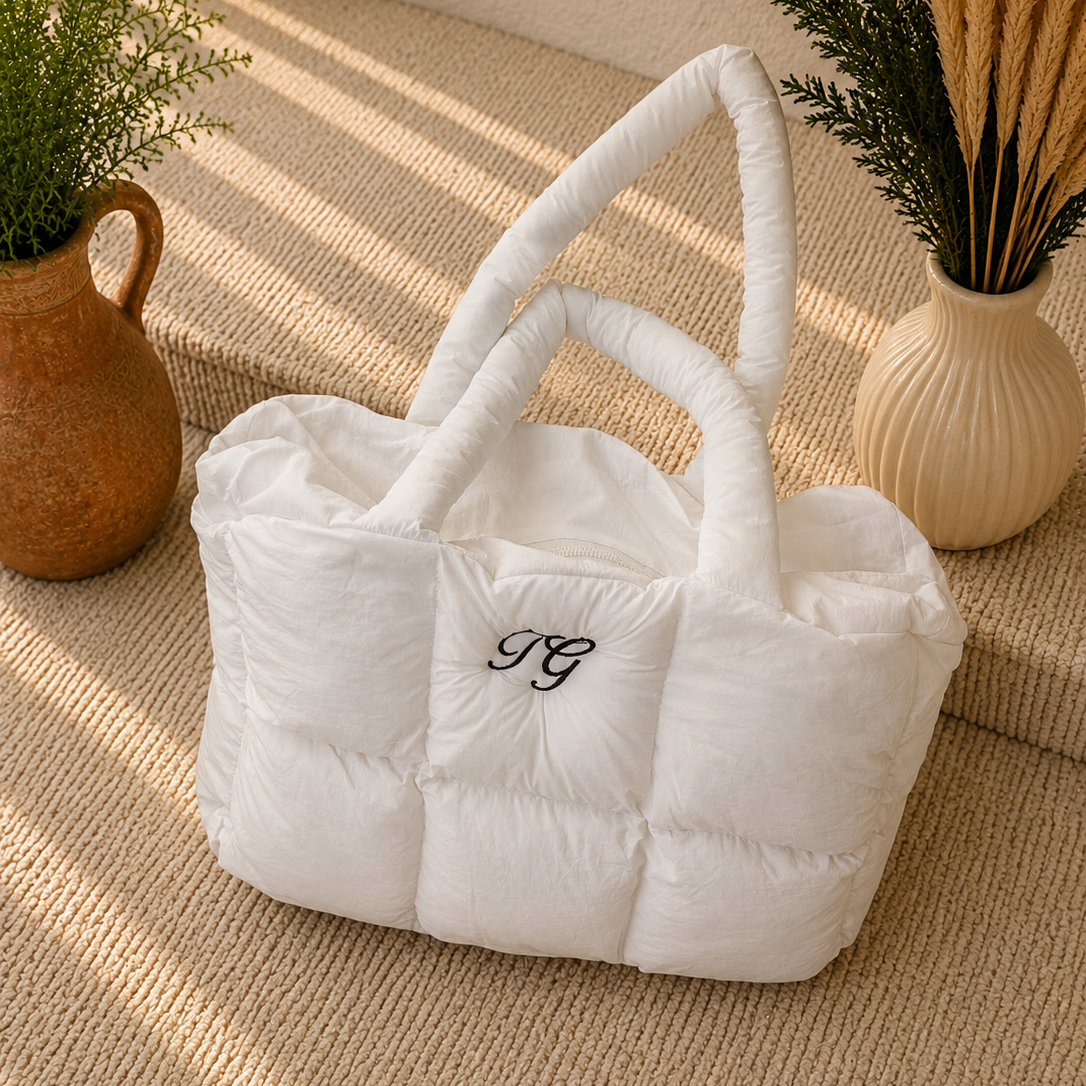 A white puffer-style tote bag with quilted cube sections and two rolled top handles, placed on a woven beige surface. The bag has a small black embroidered monogram on the front.
