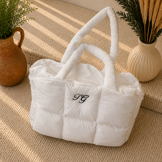 A white puffer-style tote bag with quilted cube sections and two rolled top handles, placed on a woven beige surface. The bag has a small black embroidered monogram on the front.