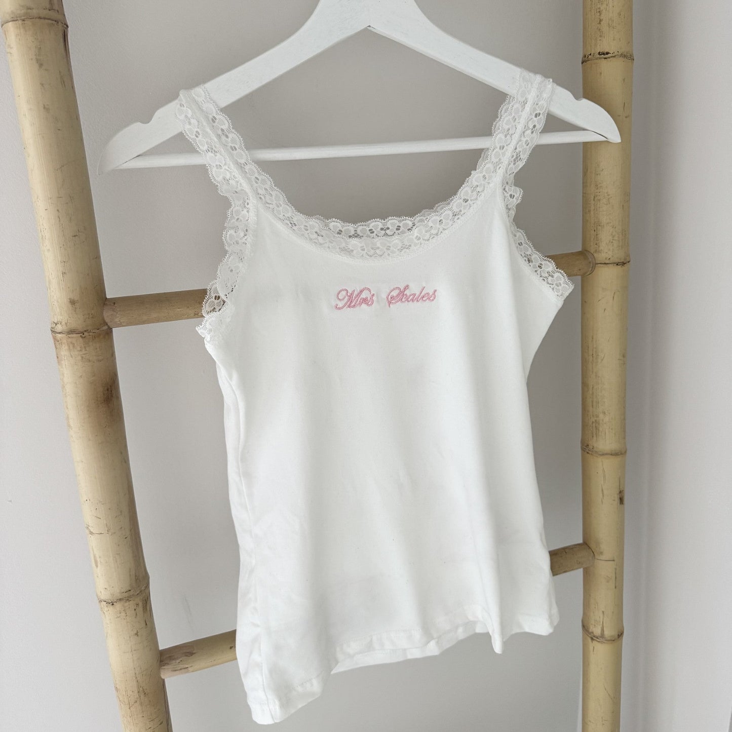 White lace-trimmed tank top with red text on a hanger against a light gray wall.