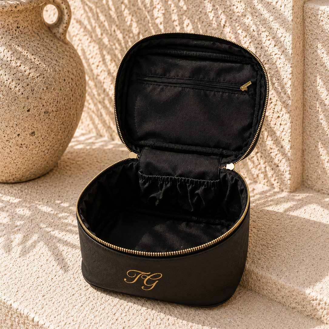 A black zippered cosmetic case with gold trim and a gold monogram logo, shown open on a textured beige stone surface with a ceramic vase and leaf shadows in the background.