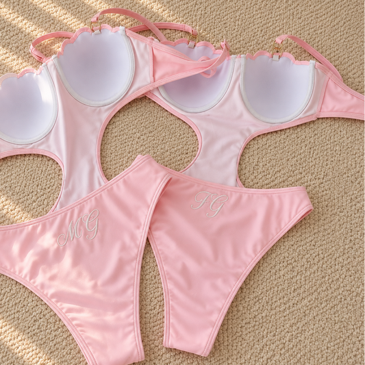 A pink two-piece bikini set laid flat on a beige textured surface. The bikini features scalloped underwire cups with white padding and thin shoulder straps, paired with matching low-rise bottoms that have a small white embroidered monogram.