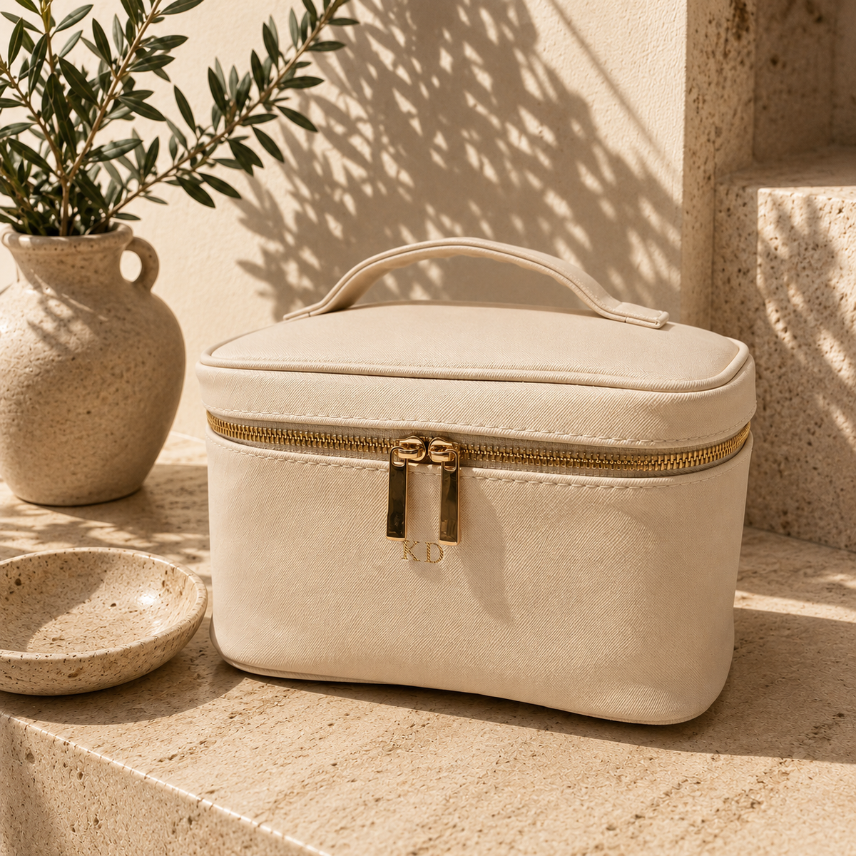 A cream-colored faux leather cosmetic bag with a gold zipper and handle, styled on a stone surface next to a ceramic vase with greenery.
