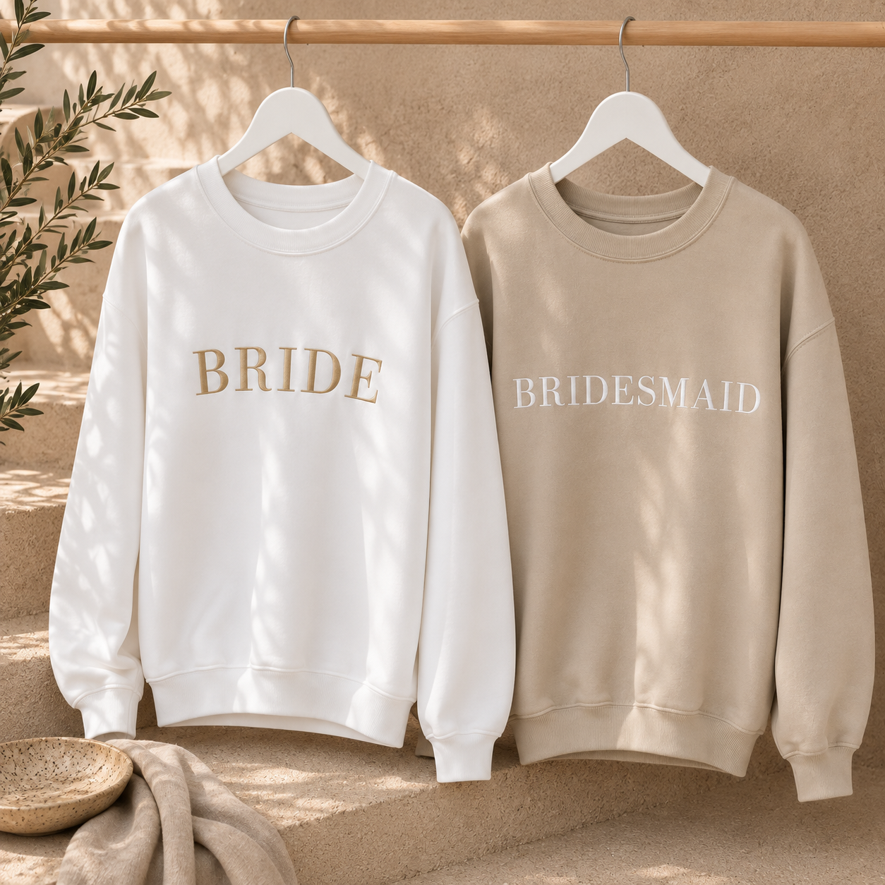 Two crewneck sweatshirts hanging on white hangers against a warm beige textured wall. The left sweatshirt is white with the word 'BRIDE' in gold lettering, and the right sweatshirt is beige with the word 'BRIDESMAID' in white lettering.