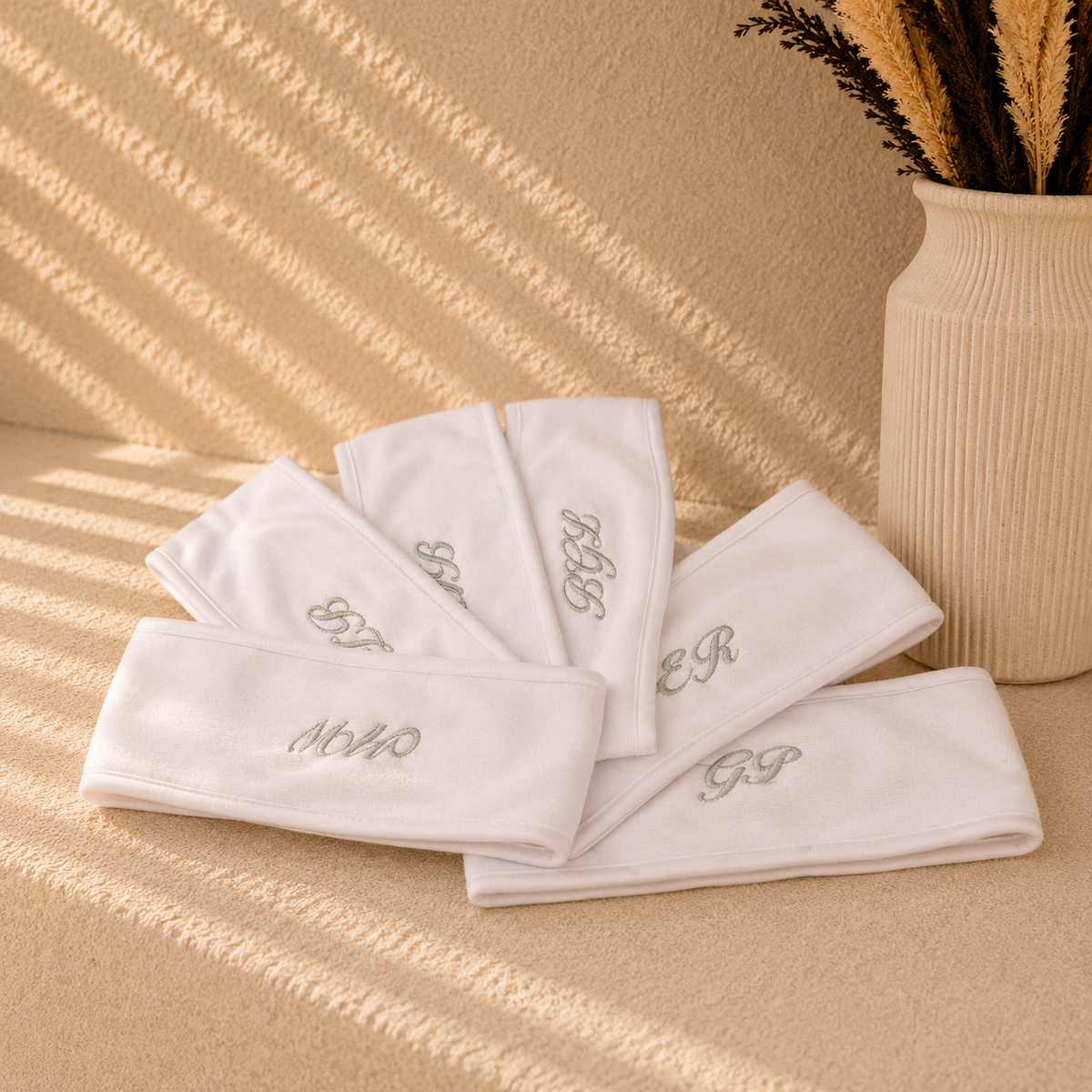 Five white cotton headbands with embroidered monogram initials, arranged on a textured beige surface with warm, dappled sunlight and a decorative vase in the background.