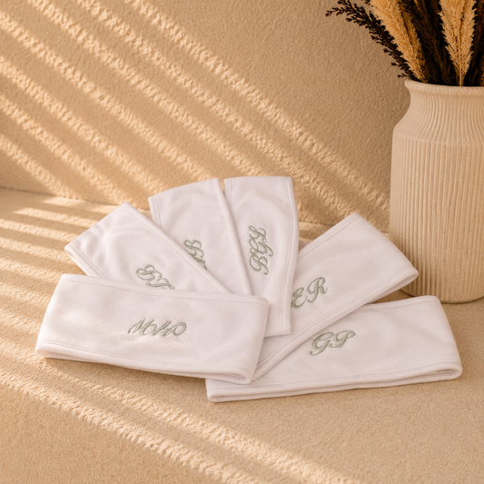 Five white cotton headbands with embroidered monogram initials, arranged on a textured beige surface with warm, dappled sunlight and a decorative vase in the background.