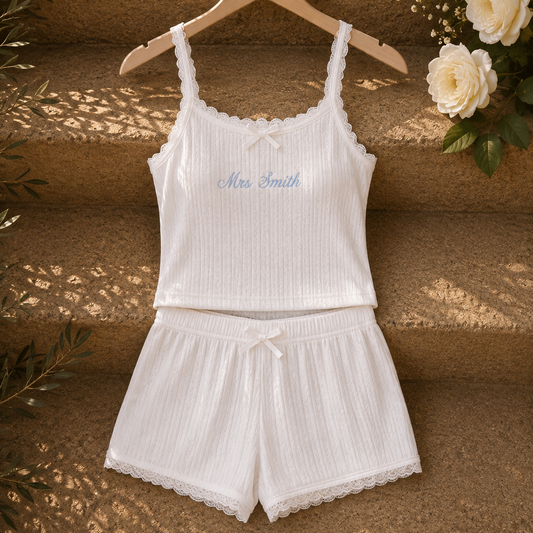 A two-piece white ribbed pajama set with a camisole top and matching shorts, hanging on a wooden hanger against a rustic stone wall with flowers and leaf shadows.