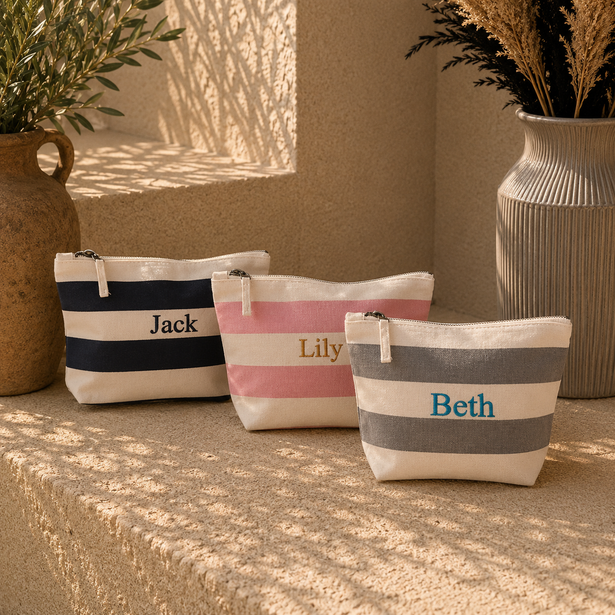 Three personalized canvas zip pouches with horizontal stripes are arranged on a textured stone surface in a warm, sunlit setting. The pouches are cream-colored with navy blue, pink, and gray stripes, respectively, and are personalized with the names Jack, Lily, and Beth.