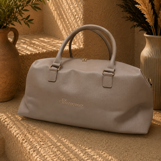 A light taupe-gray personalized duffel bag with gold hardware and the name 'Shannon' embroidered in gold on the front, styled on a textured beige stone surface with warm sunlight and decorative vases.
