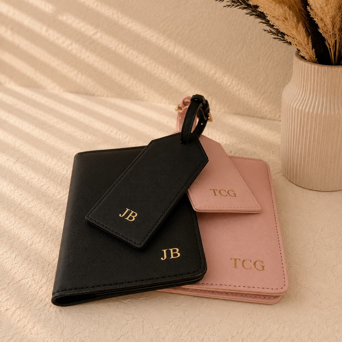 Two personalized leather luggage tags and matching card cases, one black and one dusty pink, with gold monogram initials 'JB' and 'TCG', styled on a textured beige surface with a vase in the background.