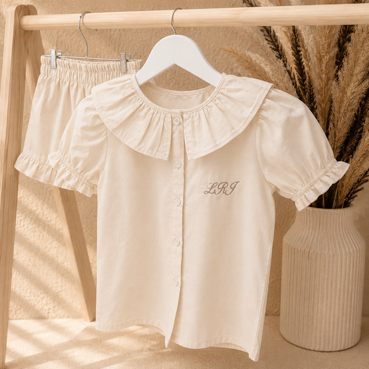 A cream-colored short-sleeve button-front blouse with a large ruffled collar and matching ruffled sleeves, hanging on a white hanger on a wooden clothing rack. A pair of matching shorts is partially visible behind it.