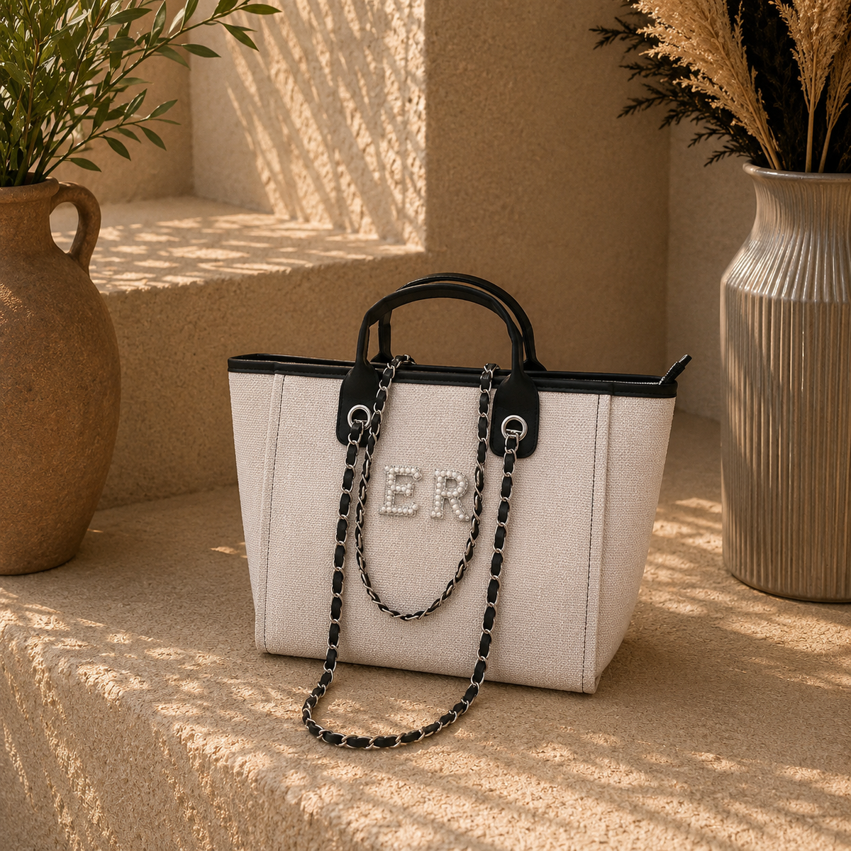 A structured cream-colored tote bag with black trim, a black-and-silver chain strap, and rhinestone-embellished lettering on the front, styled on a sunlit beige stone surface with potted plants in the background.