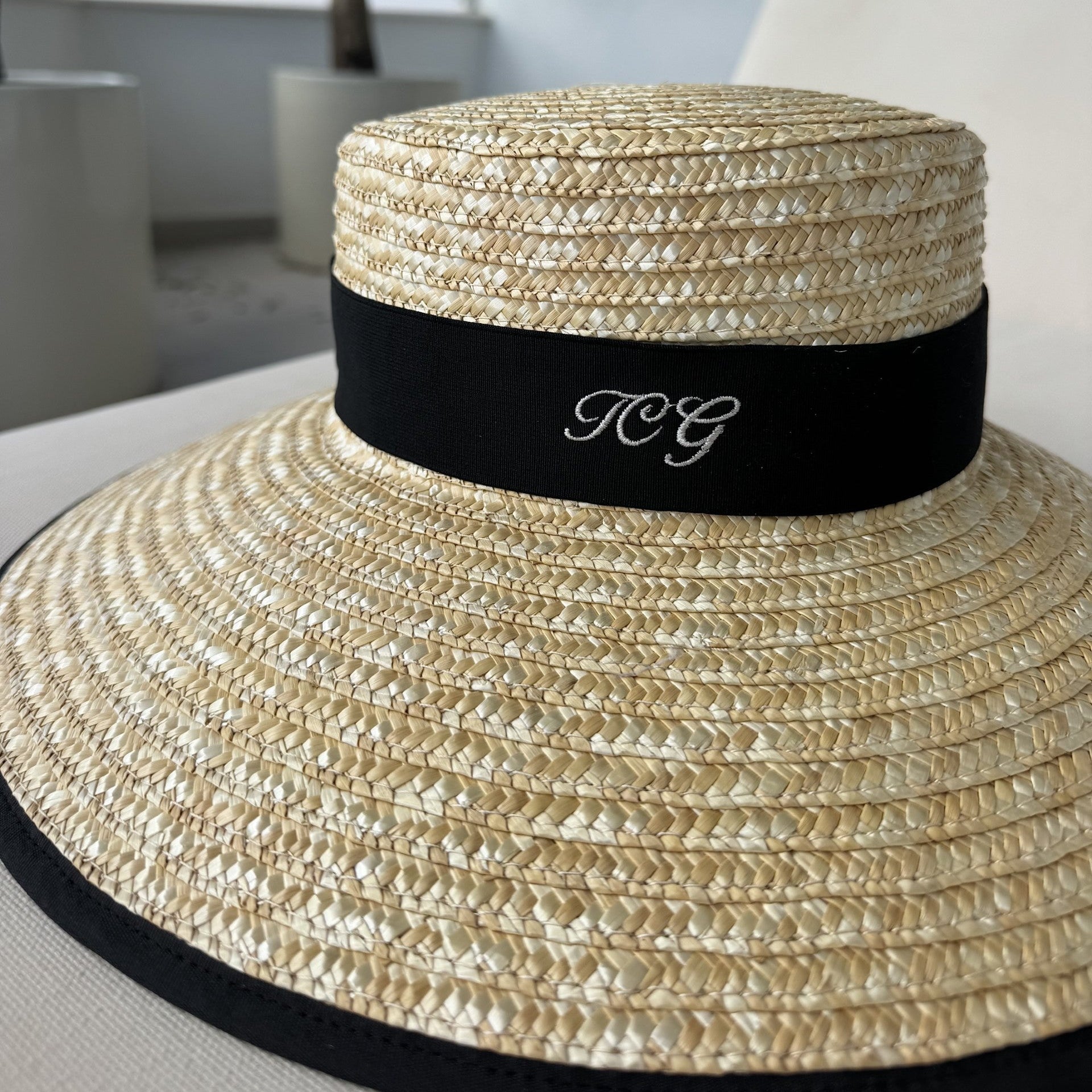 Beige straw hat with a black band featuring initials, placed on a white surface.