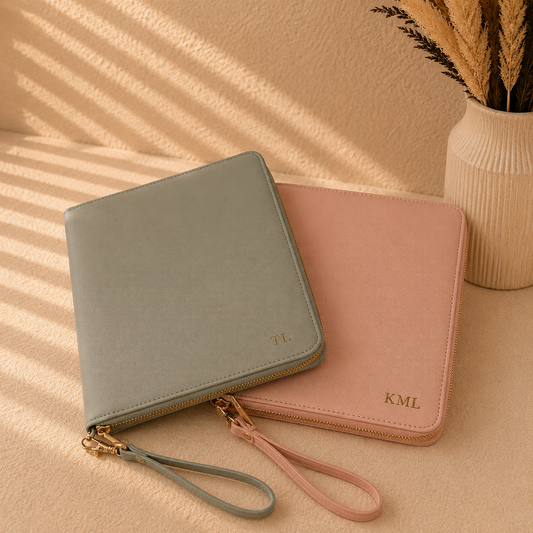 Two zip-around wallet-style organizers with wrist straps, one in muted sage green and one in dusty pink, arranged on a beige surface with decorative dried grass in a vase. Each wallet has gold-tone hardware and a small gold monogram.