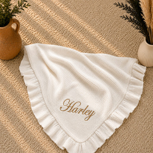 A white textured baby blanket with ruffled edges and the name 'Harley' embroidered in gold script, styled on a beige woven rug with decorative plants in vases.