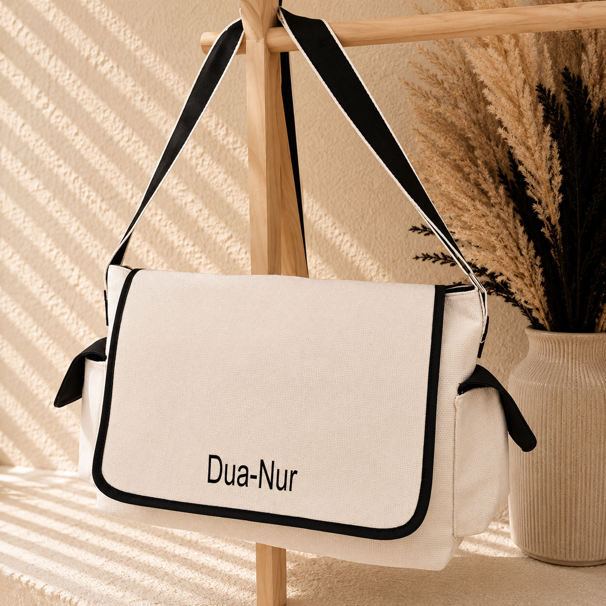A cream canvas shoulder bag with black trim and a black adjustable strap, featuring the brand name 'Dua-Nur' printed on the front. The bag is hanging from a wooden stand in a styled setting with warm sunlight and dried pampas grass in a vase.