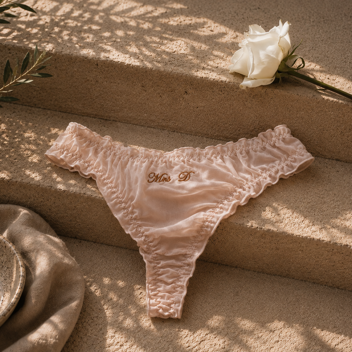 A pair of light blush pink ruffled thong underwear with gold embroidered initials 'M & T' on the front, styled on a textured beige stone surface with a white rose and a speckled bowl nearby.