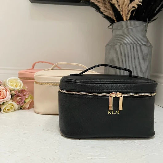 Black cosmetic bag with gold zippers and 'KLM' monogram with beige and pink cosmetic bag in the background