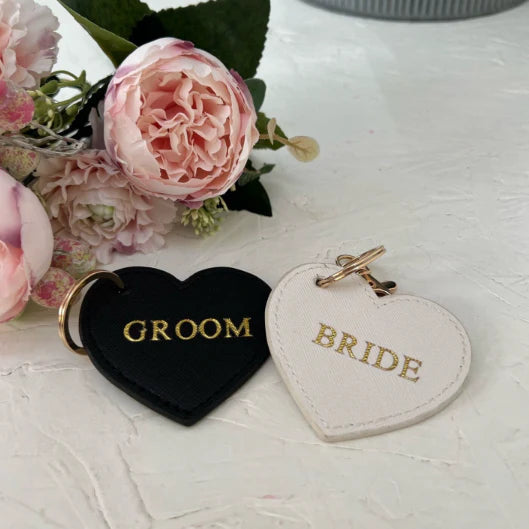 Heart-shaped 'GROOM' and 'BRIDE' keychains on a light surface with flowers in the background.