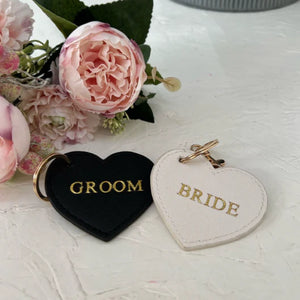 Heart-shaped 'GROOM' and 'BRIDE' keychains on a light surface with flowers in the background.