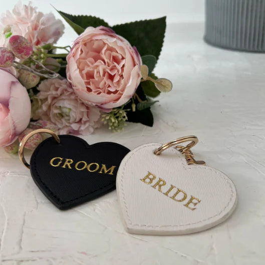 Heart-shaped 'Groom' and 'Bride' keychains on a light surface with flowers in the background.