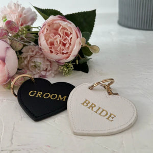 Heart-shaped 'Groom' and 'Bride' keychains on a light surface with flowers in the background.