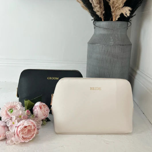 Beige 'BRIDE' pouch and black 'GROOM' pouch with flowers and a vase in the background.