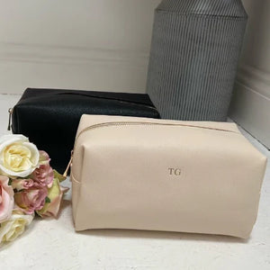 Beige toiletry bag with 'TG' initials on a light surface with flowers and a black bag in the background.
