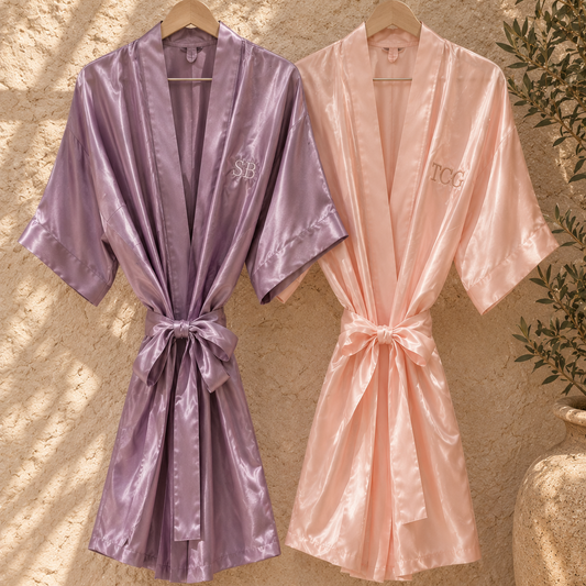 Two satin-like robes hang on wooden hangers against a textured beige wall. The left robe is lavender with a monogram reading 'SBB' on the chest, and the right robe is blush pink with a monogram reading 'TCG'.