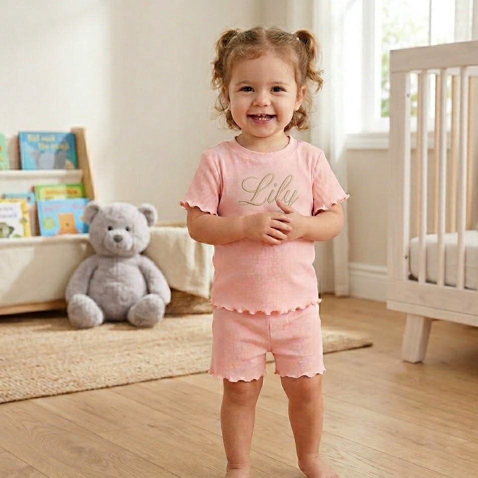 Child wearing pink set with 'Lily' on a wooden floor in a nursery.