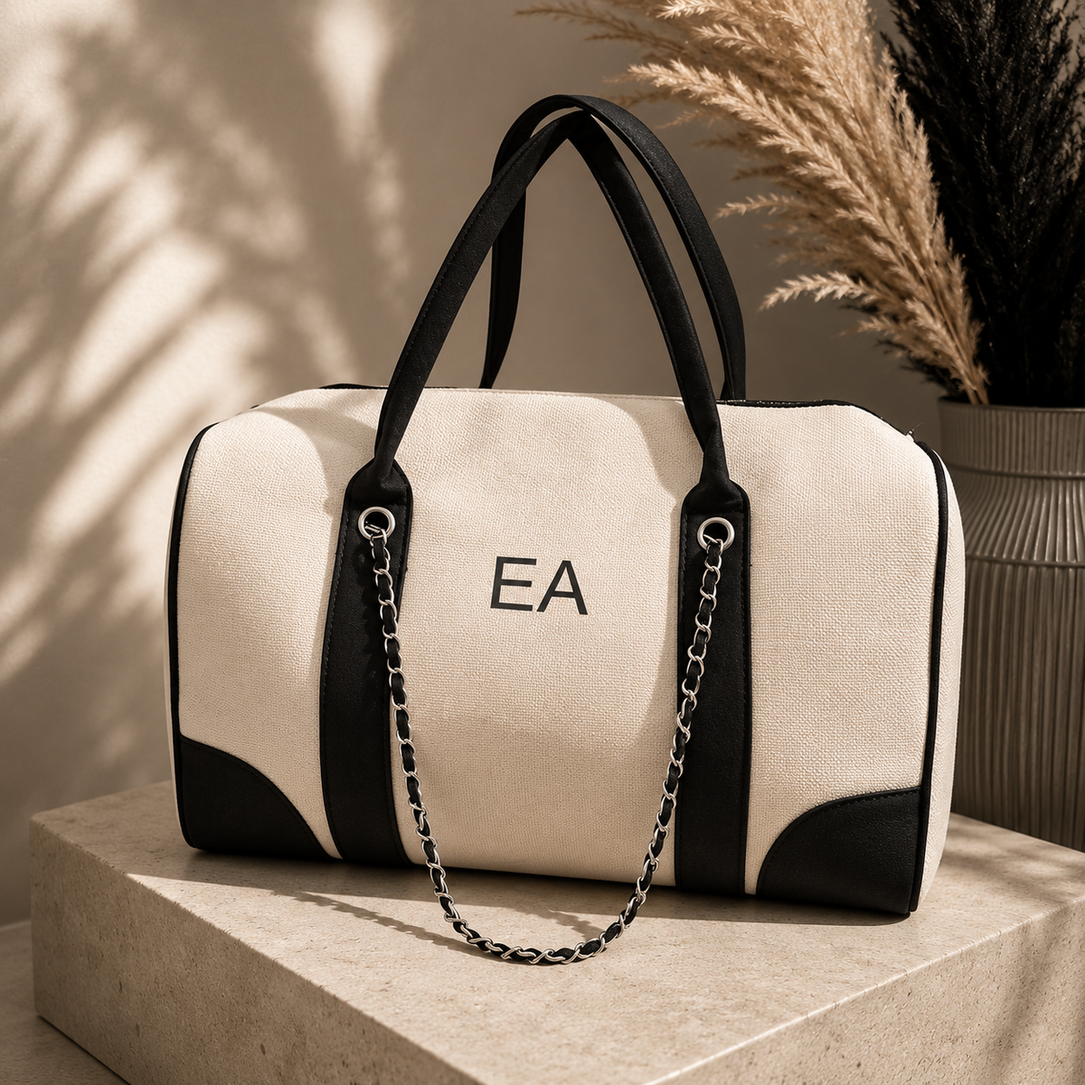 A cream canvas duffel bag with black leather trim, double top handles, and a silver chain strap, displayed on a stone pedestal with pampas grass in the background. The bag features the monogram 'EA' on the front.
