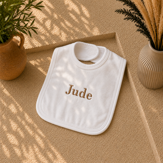 A white baby bib with the name 'Jude' embroidered in brown on the front, styled on a textured beige surface with potted plants and leaf shadows.