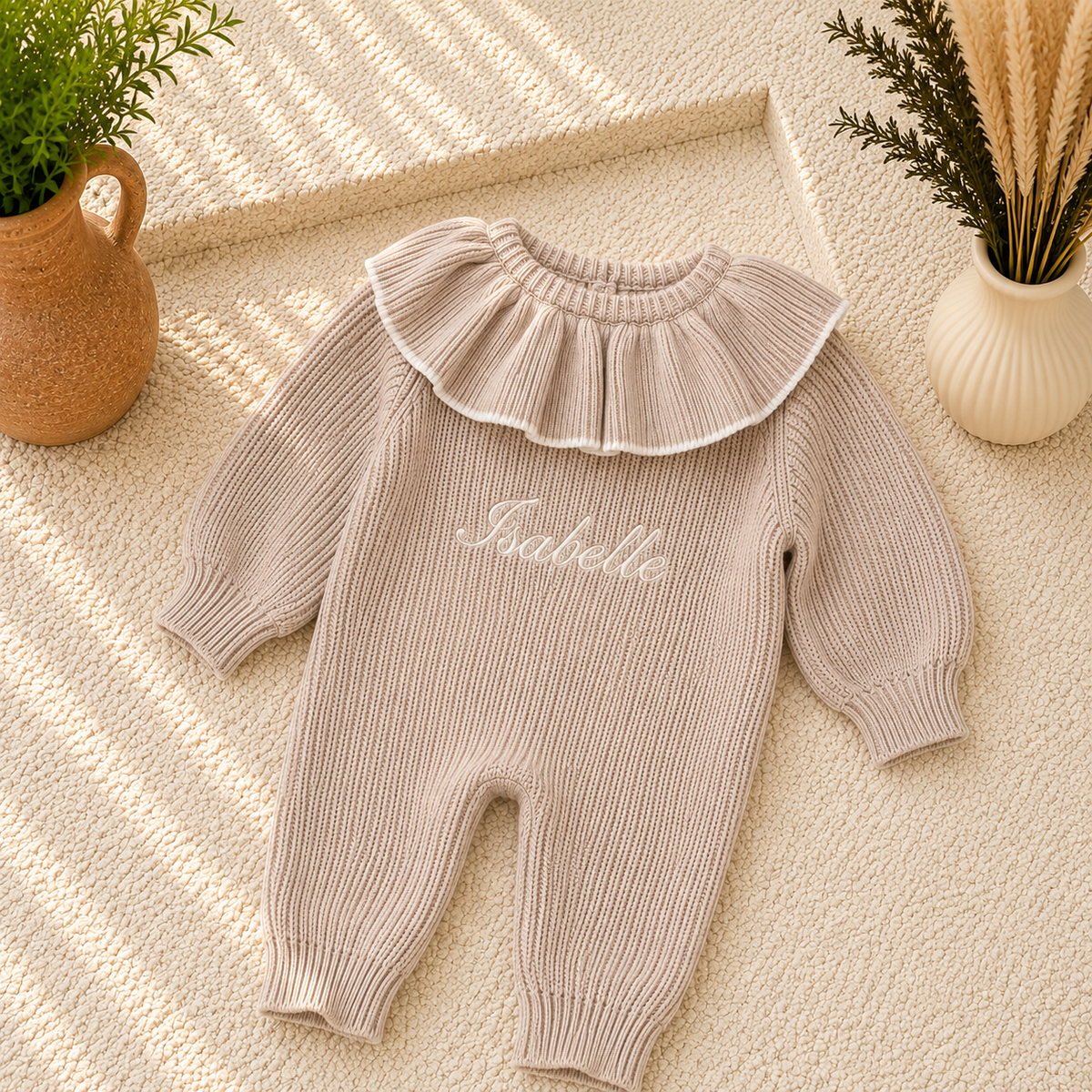 A baby knit jumpsuit in a warm taupe-beige color with a wide ruffled collar and white embroidered script reading 'Isabelle' on the chest. The jumpsuit is styled in a flat-lay composition on a textured cream rug with decorative plants in ceramic pots.