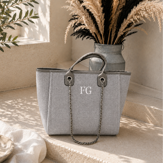 A light gray fabric tote bag with dark gray leather trim, a top handle, and a silver chain strap, placed on a beige stone surface. The bag has the letters 'FG' printed on the front.