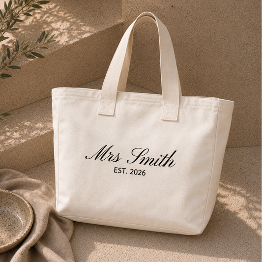 A cream canvas tote bag with black script text reading 'Mrs Smith EST. 2026' sits on a beige stone surface with a draped cloth and a small bowl nearby.