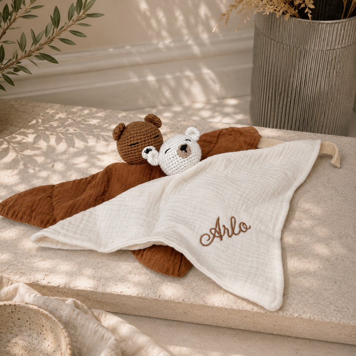 A personalized baby muslin blanket in cream white with the name 'Arlo' embroidered in brown, styled with a brown plush bear and a crocheted white bear on a beige stone surface.