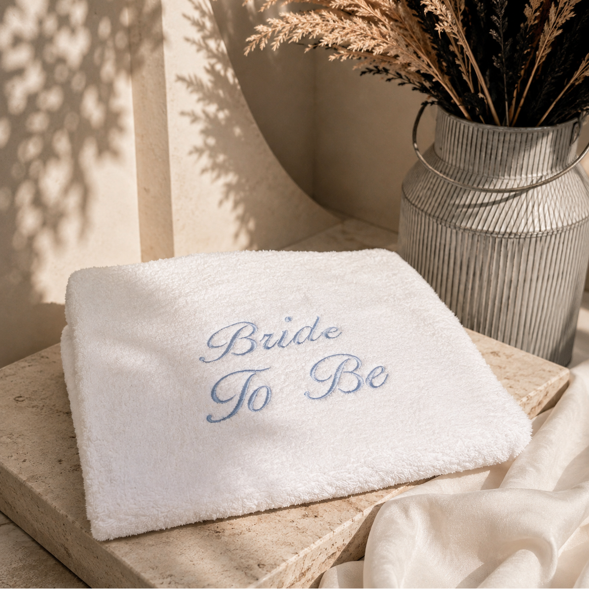 A folded white terry cloth napkin or towel with the words 'Bride To Be' embroidered in light blue script, styled on a stone surface next to a ribbed metal vase with dried grasses.