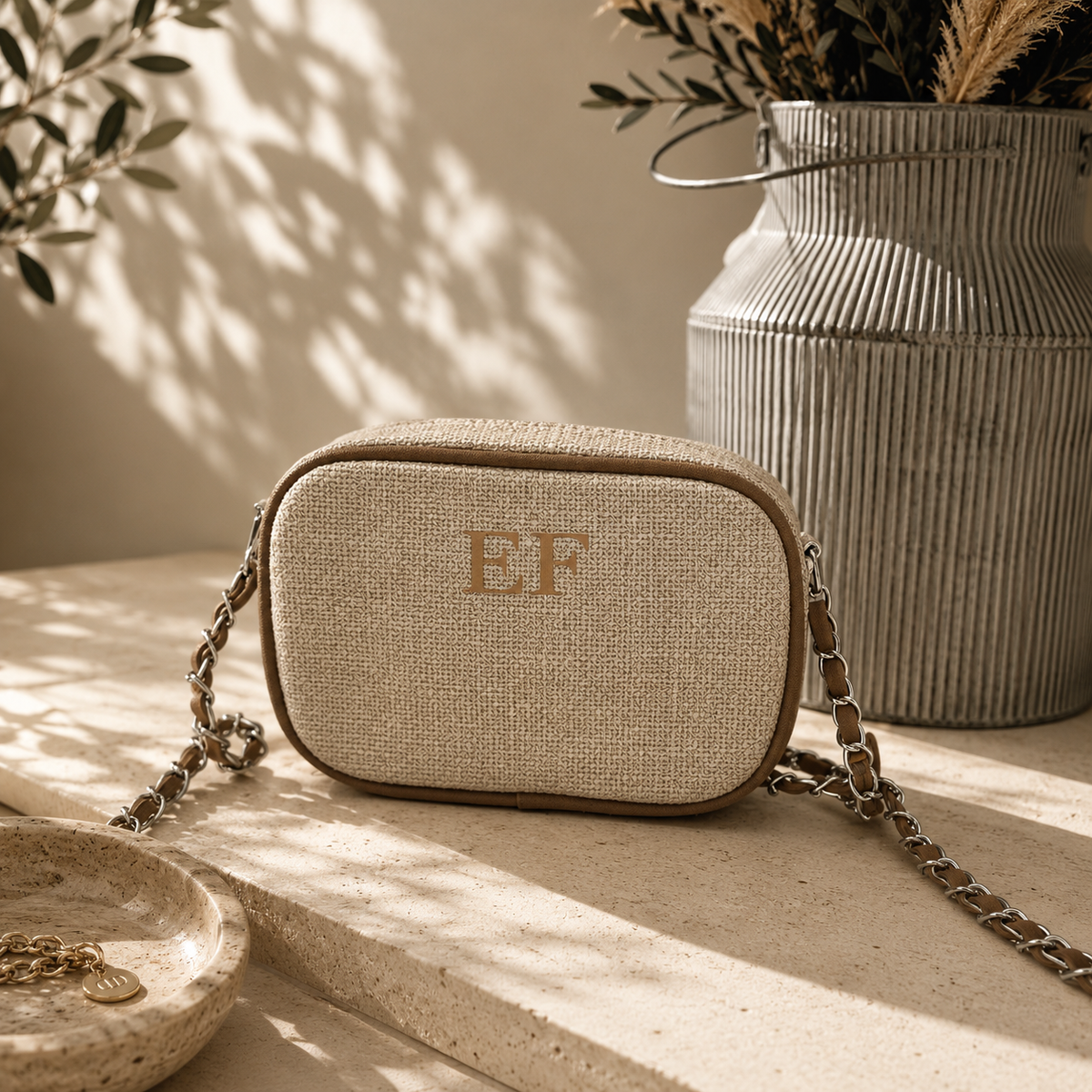 A beige woven crossbody bag with a gold-tone chain strap and monogrammed 'EF' on the front, styled on a stone ledge with warm sunlight and a ribbed metal vase in the background.