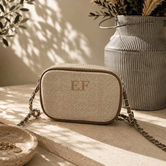 A beige woven crossbody bag with a gold-tone chain strap and monogrammed 'EF' on the front, styled on a stone ledge with warm sunlight and a ribbed metal vase in the background.
