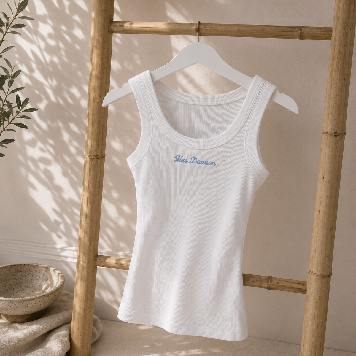 A white ribbed tank top with 'Mia Dawson' embroidered in blue script on the chest, hanging on a white hanger against a beige wall with bamboo ladder props.
