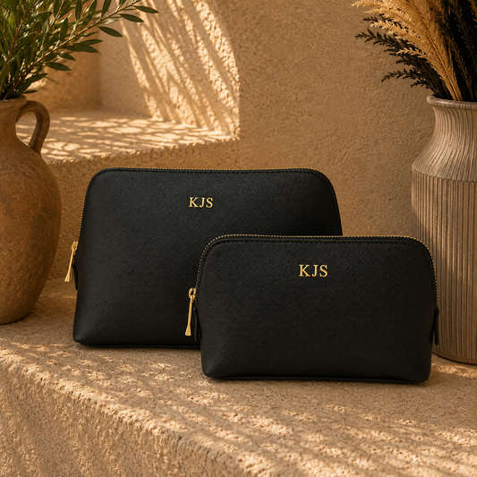 Two black textured zippered cosmetic pouches with gold zippers and gold 'KJS' lettering, displayed on a sunlit stone ledge with decorative vases in the background.