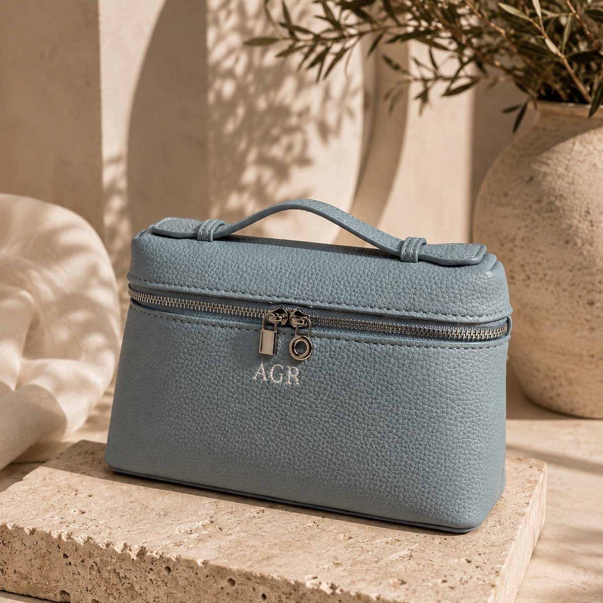A personalized light blue pebbled leather cosmetic bag with a top handle, silver zipper, and two padlocks, featuring the monogram 'AGR' on the front. The bag is styled on a textured stone pedestal with warm sunlight and shadows.