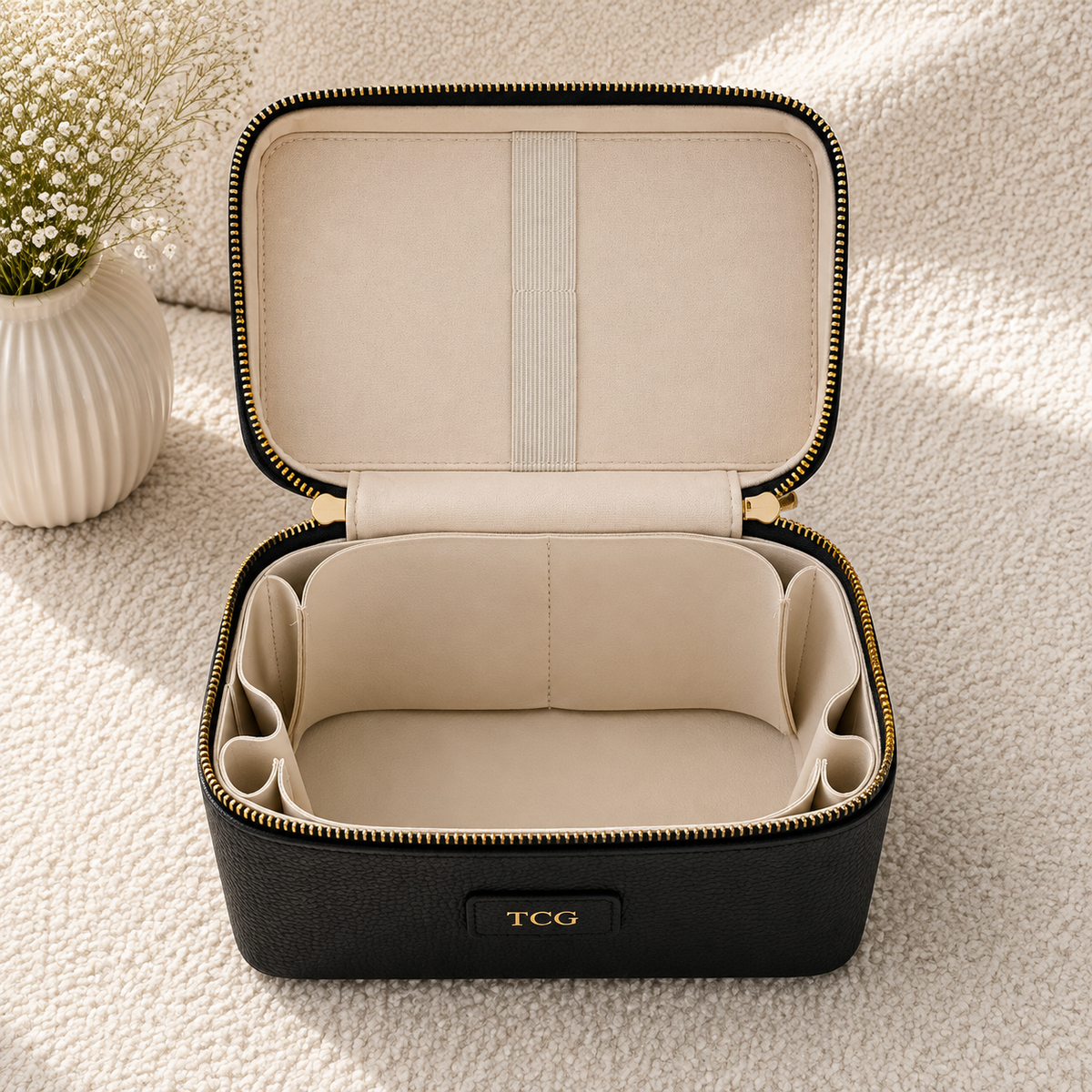An open black zippered cosmetic case with a beige interior, shown on a soft cream textured surface. The case has gold-tone zipper trim, side compartments, and a front label reading TCG.