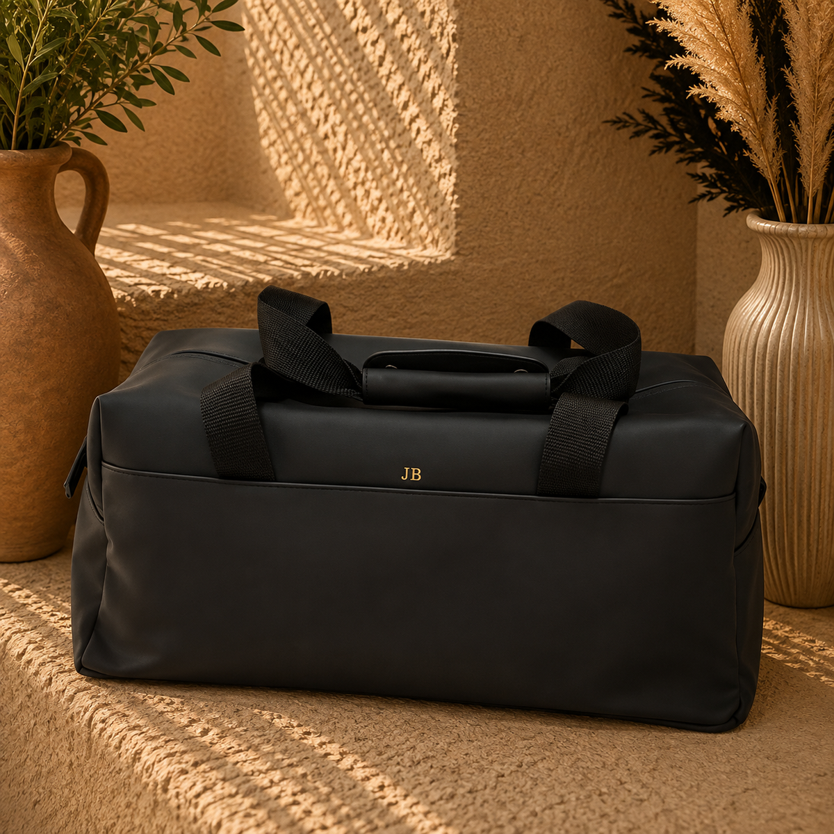 A black duffel bag with top handles sits on a woven rug in a warm, styled interior setting with potted plants and textured walls. The bag has a small gold 'JB' monogram on the front.