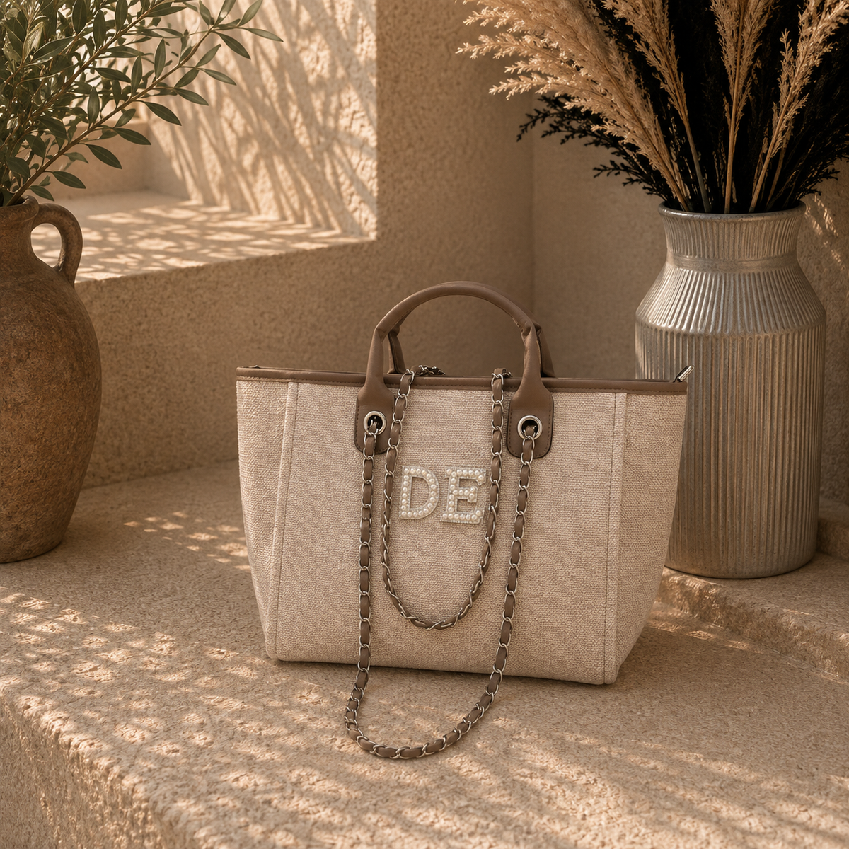 A structured beige tote bag with brown leather handles and trim, featuring a chain strap and a rhinestone-embellished 'DE' logo on the front. The bag is styled on a textured stone ledge between two decorative vases in warm, sunlit tones.