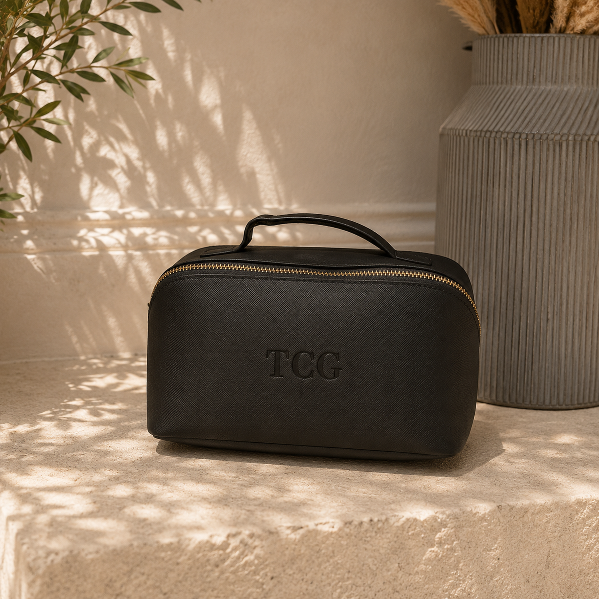 A black textured leather cosmetic case with a top handle and gold zipper, featuring an embossed 'TCC' logo, set on a stone ledge with warm natural light and a ribbed ceramic vase in the background.