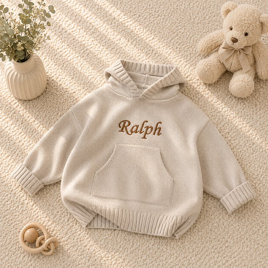 A cream-colored knit baby hoodie with 'Ralph' embroidered in brown on the chest, styled in a flat lay with a teddy bear, a small vase of flowers, and a wooden pacifier on a textured beige blanket.