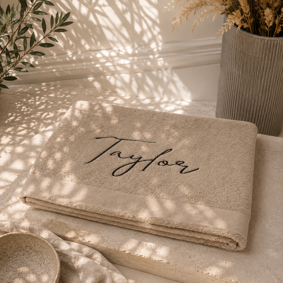 A folded beige towel with the name 'Taylor' embroidered in black script across the center, styled on a stone surface with warm natural shadows, dried grass in a vase, and a small bowl nearby.