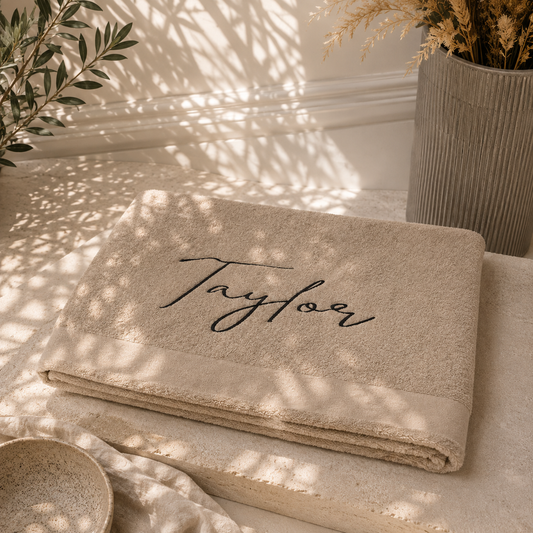 A folded beige towel with the name 'Taylor' embroidered in black script across the center, styled on a stone surface with warm natural shadows, dried grass in a vase, and a small bowl nearby.